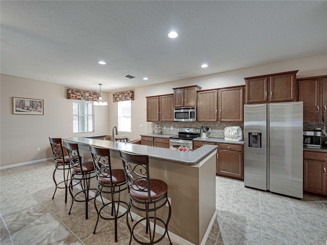 kitchen with tasteful backsplash, appliances with stainless steel finishes, decorative light fixtures, and a kitchen island with sink