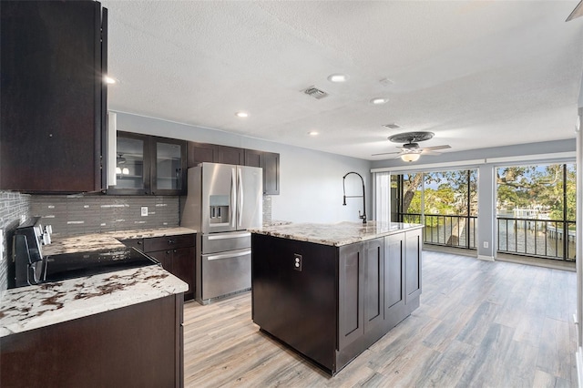 kitchen featuring an island with sink, stainless steel fridge with ice dispenser, light wood-type flooring, black range, and light stone countertops