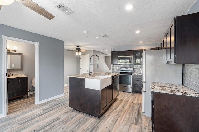 kitchen with decorative backsplash, appliances with stainless steel finishes, a textured ceiling, a kitchen island with sink, and light hardwood / wood-style flooring