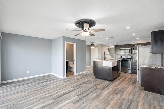 kitchen featuring decorative backsplash, stainless steel appliances, a kitchen island with sink, ceiling fan, and hardwood / wood-style flooring