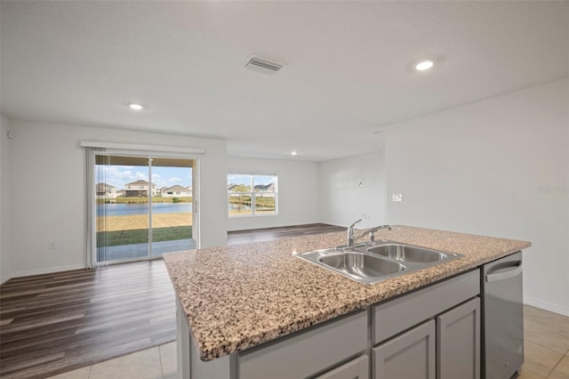 kitchen featuring a center island with sink, sink, light stone counters, stainless steel dishwasher, and light wood-type flooring