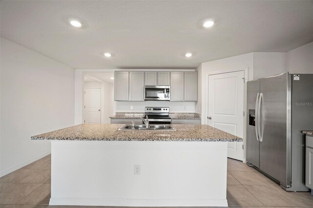 kitchen with gray cabinetry, appliances with stainless steel finishes, light stone counters, and a center island with sink