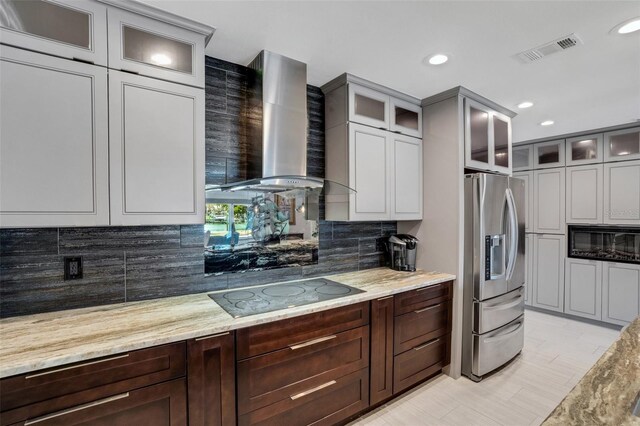 kitchen with wall chimney exhaust hood, decorative backsplash, black electric cooktop, light stone counters, and stainless steel fridge with ice dispenser