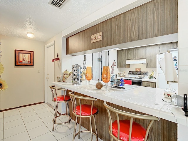 kitchen with a textured ceiling, white appliances, light tile patterned floors, tile counters, and a breakfast bar area