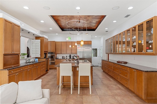 kitchen featuring a breakfast bar, stainless steel built in fridge, a tray ceiling, a kitchen island with sink, and pendant lighting