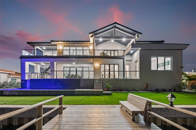 back of property at dusk featuring stucco siding, a balcony, a lawn, and stairway