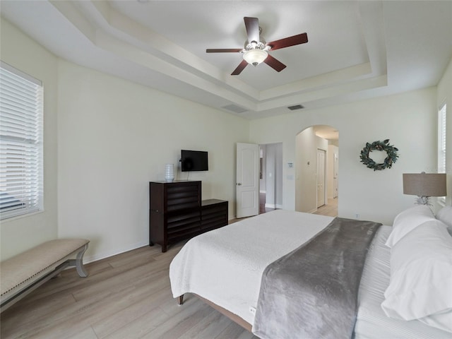 bedroom featuring multiple windows, light hardwood / wood-style flooring, ceiling fan, and a tray ceiling