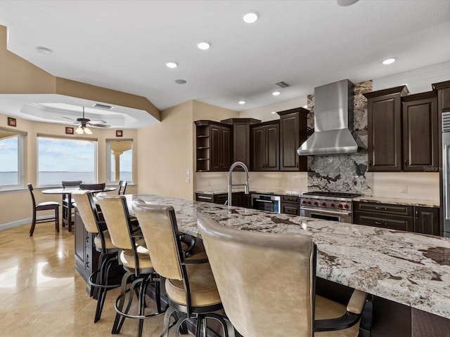 kitchen with ceiling fan, wall chimney range hood, a kitchen breakfast bar, light stone counters, and high end range