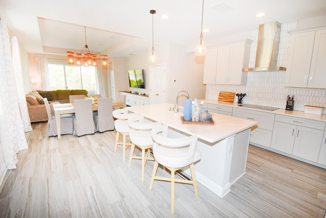 kitchen with light hardwood / wood-style floors, black electric stovetop, wall chimney exhaust hood, an island with sink, and a breakfast bar