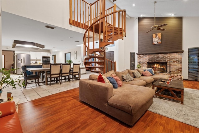 living room with a brick fireplace, light hardwood / wood-style floors, ceiling fan, and a towering ceiling