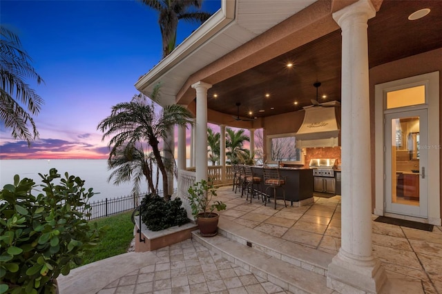 patio terrace at dusk with a bar, exterior kitchen, ceiling fan, and area for grilling