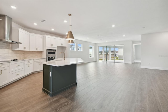 kitchen with pendant lighting, wall chimney range hood, white cabinetry, a kitchen island with sink, and stainless steel double oven