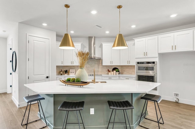 kitchen with wall chimney exhaust hood, baseboards, stainless steel double oven, light wood finished floors, and tasteful backsplash
