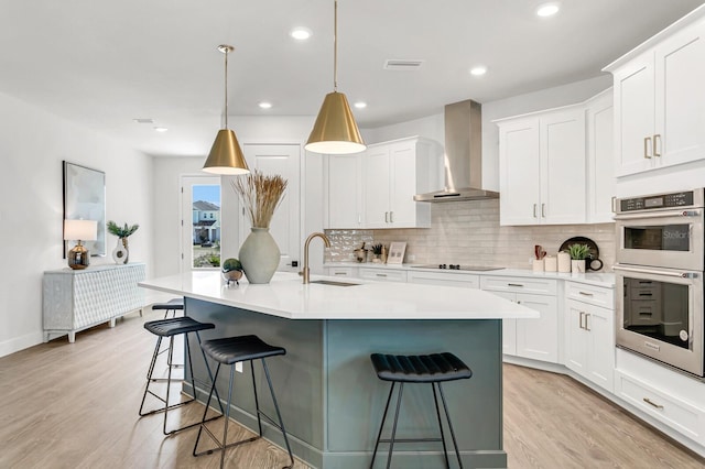 kitchen with wall chimney range hood, stainless steel double oven, a sink, black electric cooktop, and light wood-type flooring