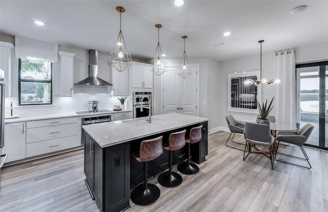 kitchen with stainless steel double oven, wall chimney range hood, hanging light fixtures, and an island with sink