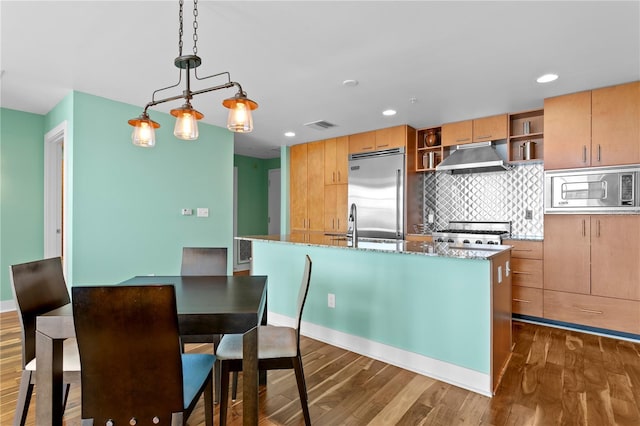 kitchen with stone counters, dark hardwood / wood-style flooring, built in appliances, decorative light fixtures, and a center island with sink