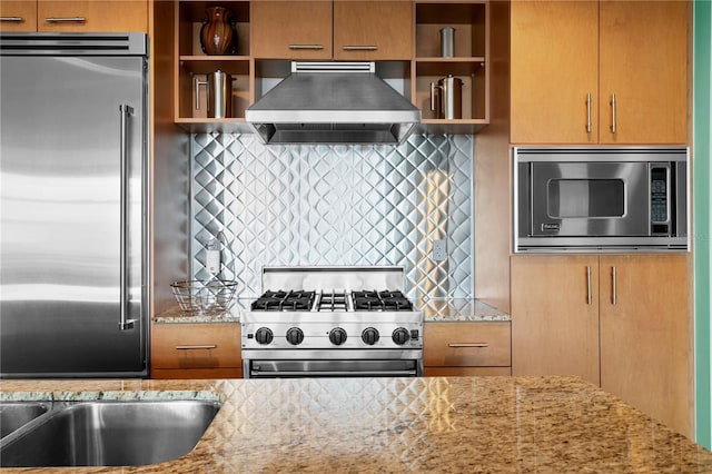 kitchen featuring built in appliances, light stone counters, wall chimney exhaust hood, and backsplash