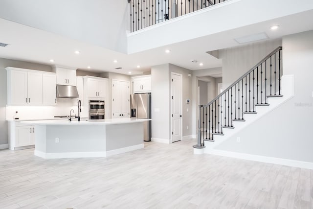 kitchen with stainless steel appliances, white cabinets, a kitchen island with sink, light hardwood / wood-style flooring, and a high ceiling