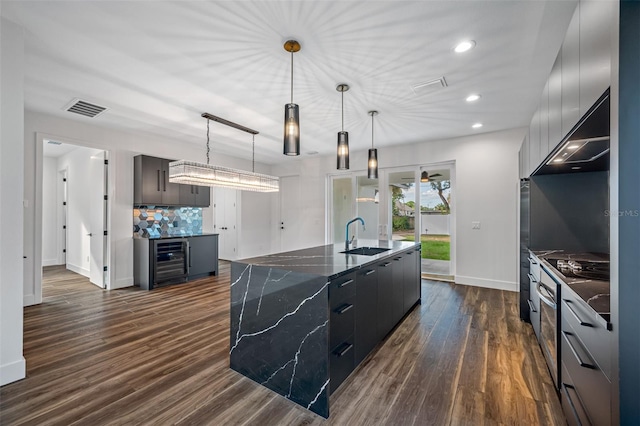 kitchen featuring dark wood-type flooring, sink, an island with sink, decorative light fixtures, and beverage cooler