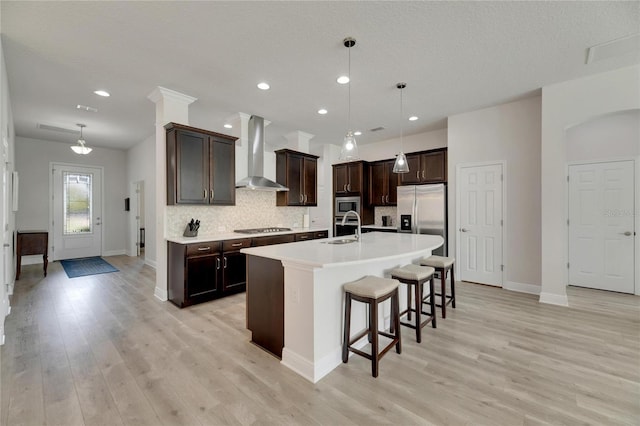 kitchen with sink, wall chimney range hood, decorative light fixtures, a kitchen island with sink, and appliances with stainless steel finishes