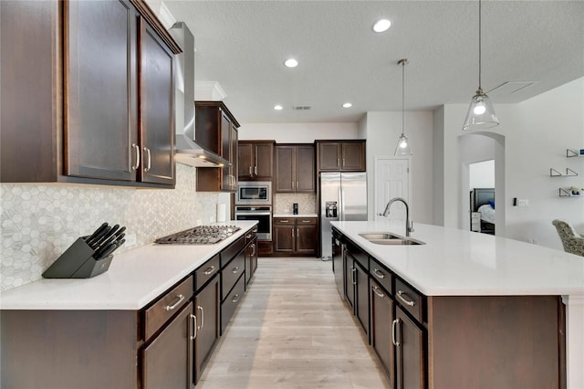 kitchen with sink, wall chimney range hood, decorative light fixtures, appliances with stainless steel finishes, and light wood-type flooring