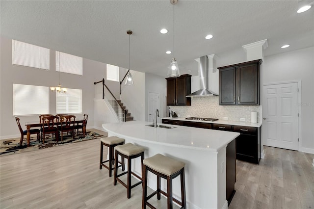 kitchen with sink, hanging light fixtures, wall chimney range hood, stainless steel gas cooktop, and an island with sink