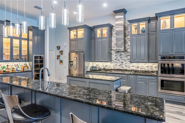 kitchen featuring hanging light fixtures, dark stone countertops, appliances with stainless steel finishes, a kitchen island with sink, and wall chimney range hood