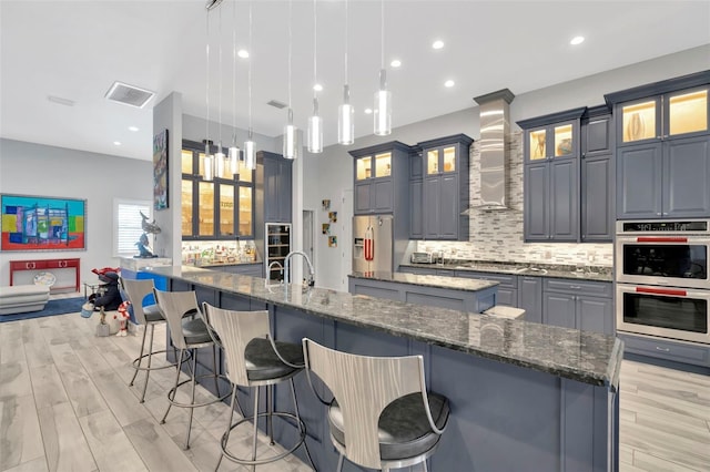 kitchen with appliances with stainless steel finishes, decorative light fixtures, dark stone counters, a large island with sink, and wall chimney range hood