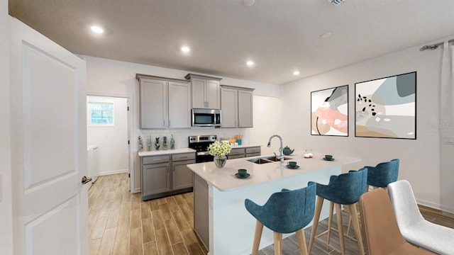 kitchen with sink, an island with sink, gray cabinetry, a breakfast bar, and appliances with stainless steel finishes