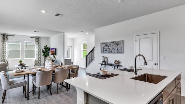 kitchen with a textured ceiling, sink, a kitchen island with sink, stainless steel dishwasher, and light wood-type flooring