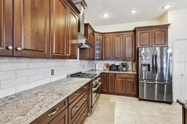 kitchen with light stone countertops, wall chimney exhaust hood, stainless steel appliances, backsplash, and a textured ceiling