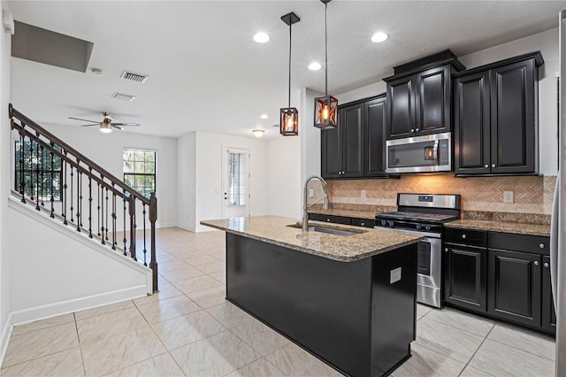 kitchen with light stone counters, stainless steel appliances, a kitchen island with sink, sink, and pendant lighting