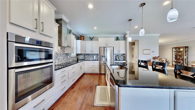 kitchen with appliances with stainless steel finishes, sink, white cabinets, hanging light fixtures, and wall chimney range hood