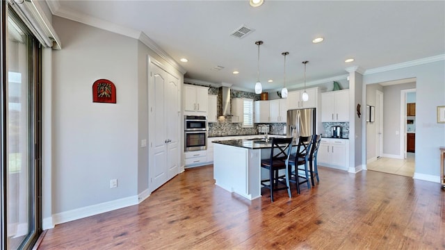 kitchen with white cabinetry, hanging light fixtures, wall chimney range hood, stainless steel appliances, and a center island with sink