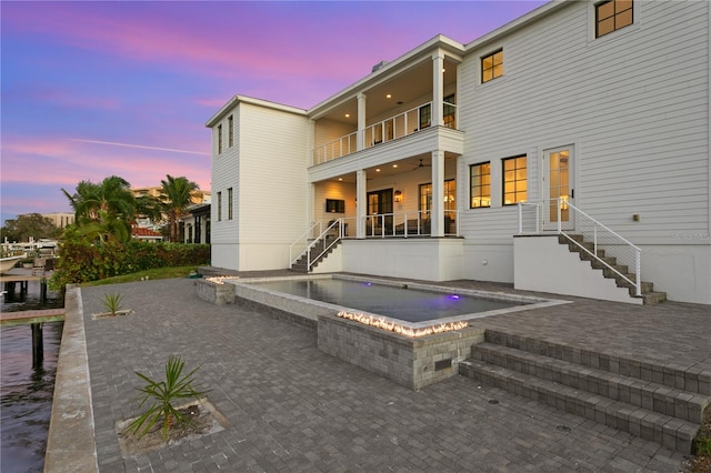 back house at dusk featuring a swimming pool with hot tub, a balcony, and a patio