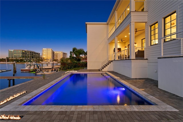 pool at dusk with a dock, ceiling fan, a patio area, and a water view