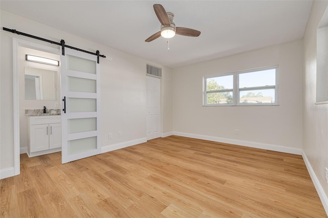 unfurnished bedroom with light hardwood / wood-style flooring, sink, a barn door, ensuite bathroom, and ceiling fan