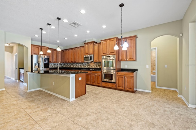 kitchen with tasteful backsplash, appliances with stainless steel finishes, a center island with sink, and decorative light fixtures