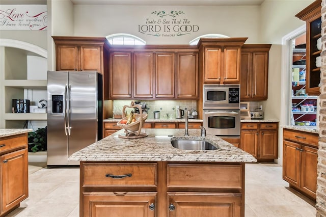 kitchen with a center island with sink, light stone counters, brown cabinets, stainless steel appliances, and a sink