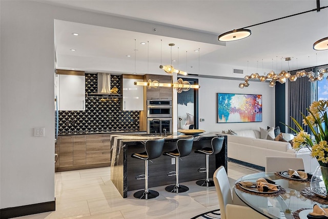 kitchen featuring wall chimney range hood, backsplash, an island with sink, pendant lighting, and a breakfast bar