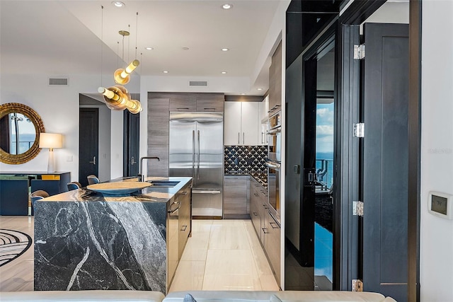 kitchen with sink, dark stone countertops, an island with sink, pendant lighting, and stainless steel built in fridge