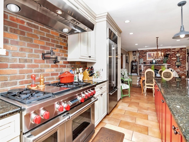 kitchen featuring high end appliances, dark stone counters, ventilation hood, hanging light fixtures, and brick wall