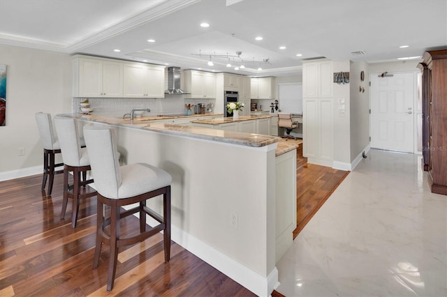 kitchen with kitchen peninsula, wall chimney exhaust hood, crown molding, light hardwood / wood-style flooring, and white cabinetry