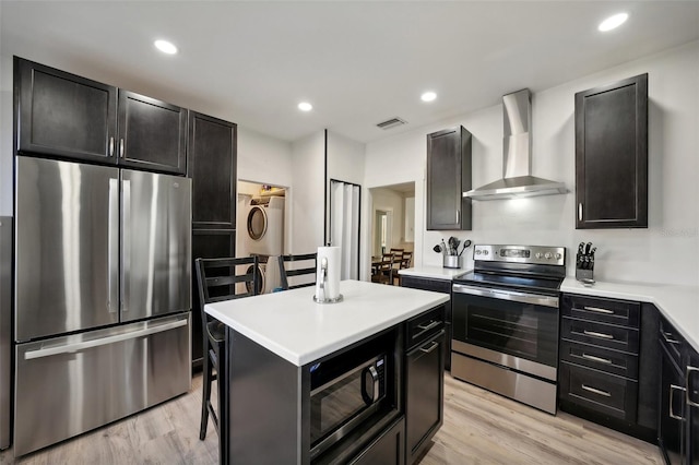 kitchen featuring a center island, wall chimney exhaust hood, stainless steel appliances, stacked washer and dryer, and light wood-type flooring