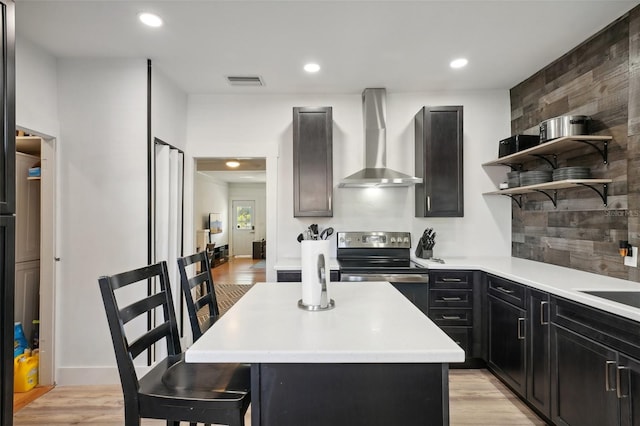 kitchen featuring stainless steel electric range, a center island, wall chimney exhaust hood, light wood-type flooring, and a kitchen bar