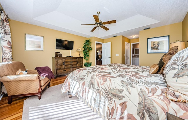 bedroom featuring a raised ceiling, ceiling fan, a textured ceiling, connected bathroom, and light hardwood / wood-style floors