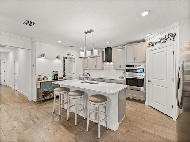 kitchen with sink, stainless steel appliances, wall chimney range hood, decorative light fixtures, and a center island with sink