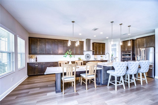 kitchen with pendant lighting, stainless steel appliances, light hardwood / wood-style flooring, and wall chimney exhaust hood