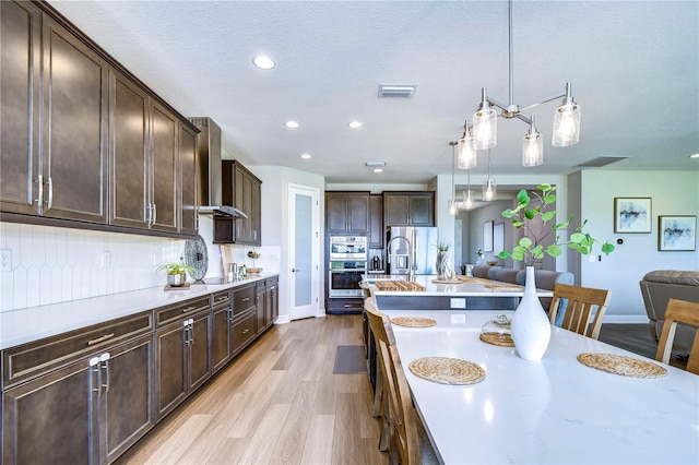 kitchen featuring dark brown cabinetry, stainless steel appliances, wall chimney range hood, light hardwood / wood-style flooring, and hanging light fixtures