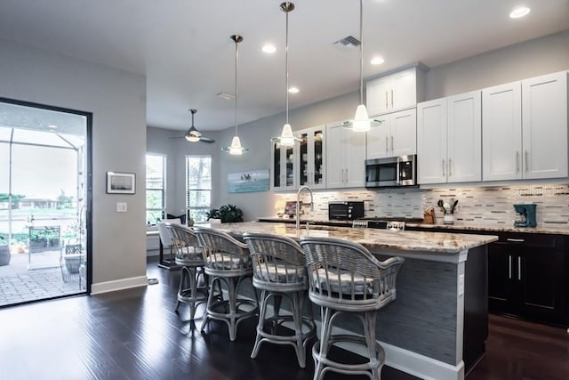 kitchen with hanging light fixtures, light stone counters, dark hardwood / wood-style floors, an island with sink, and white cabinets